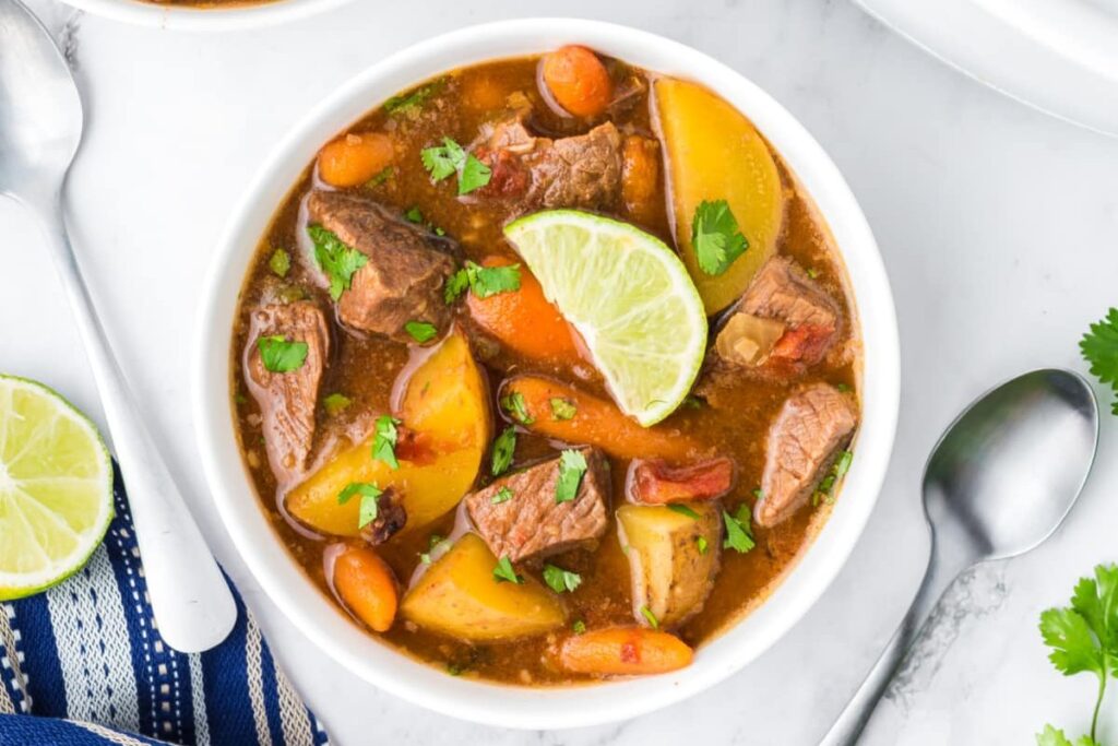 A bowl of beef stew with potatoes, carrots, and garnished with a lime slice and chopped cilantro. A spoon and a slice of lime are beside the bowl.