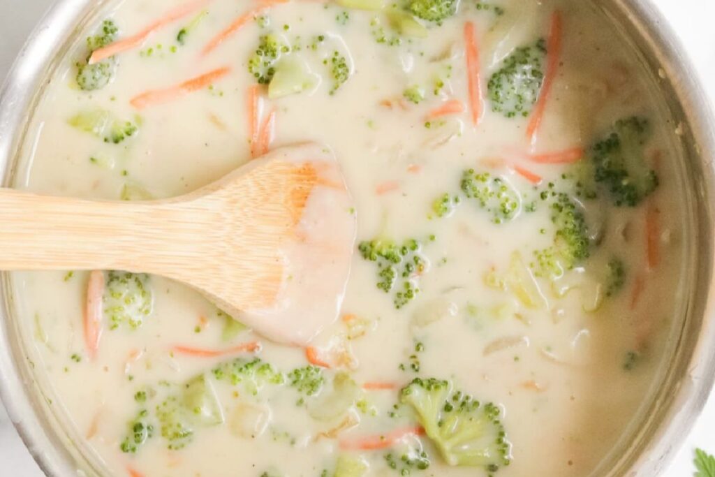 A pot of creamy broccoli and cheese soup, perfect for lunch, featuring visible chunks of broccoli, shredded carrots, and a wooden spoon stirring the mixture.